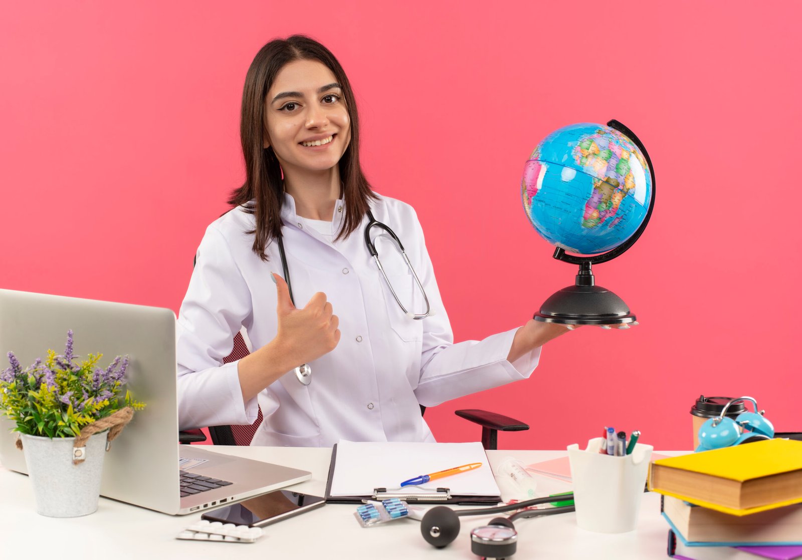 young woman doctor in white coat with stethoscope around her neck holding globe looking at camera with smile on face showing thumbs up sitting at the table with laptop over pink background