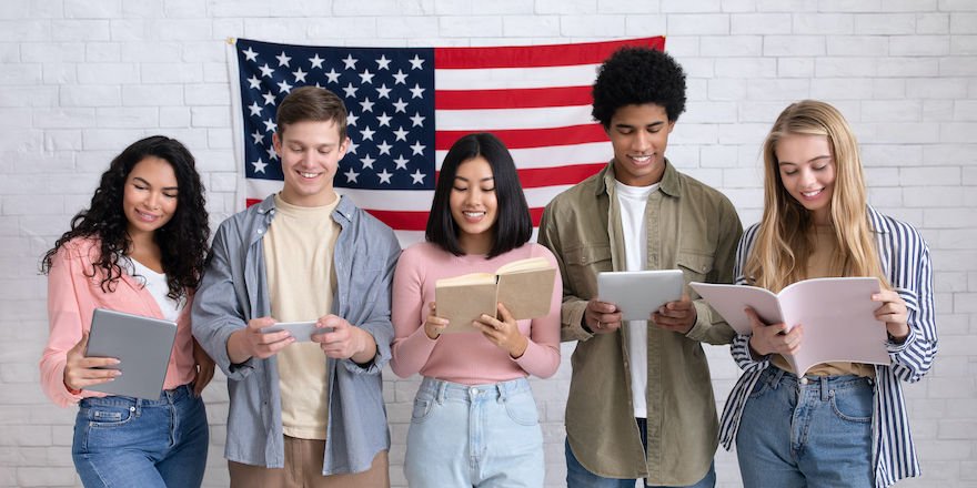 Modern study in USA, language learning and student exchange program. Cheerful busy young international people reading books and gadgets on white wall background with large flag of America, studio shot